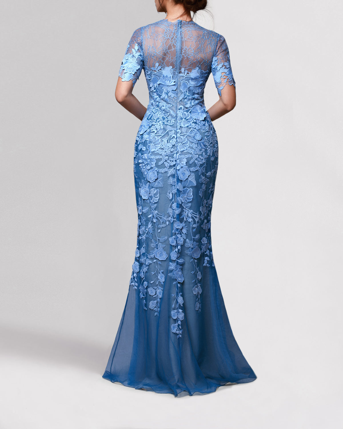 The back of a long blue evening dress featuring lace shoulders, an embroidered guipure bodice and short sleeves, continuing all the way down into a sheer skirt.