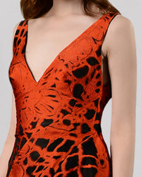 A close-up of a long evening dress in orange and black jacquard fabric featuring a V-neckline and an asymmetrical hemline.