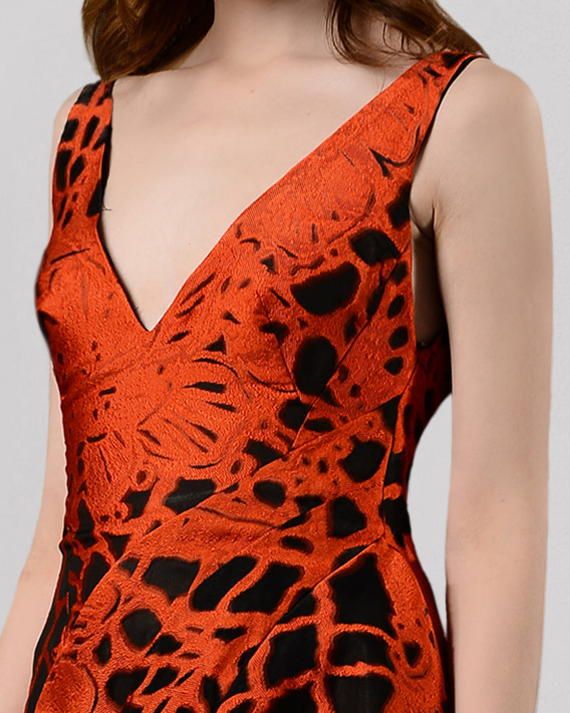 A close-up of a long evening dress in orange and black jacquard fabric featuring a V-neckline and an asymmetrical hemline.