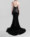 The back of a halter-neck fitted black evening gown in lace and rose patterned sequins fabric, featuring an open back and a sheer floor-length skirt with a flowing train.
