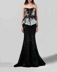 An evening wear style featuring a strapless sliver top with black lace embellishments paired with a long black mermaid cut skirt in a shimmery mesh fabric.